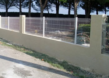 Mur bas en crépi couleur sable surmonté d'une clôture en grillage blanc rigide avec piliers en béton. Bord de chemin.