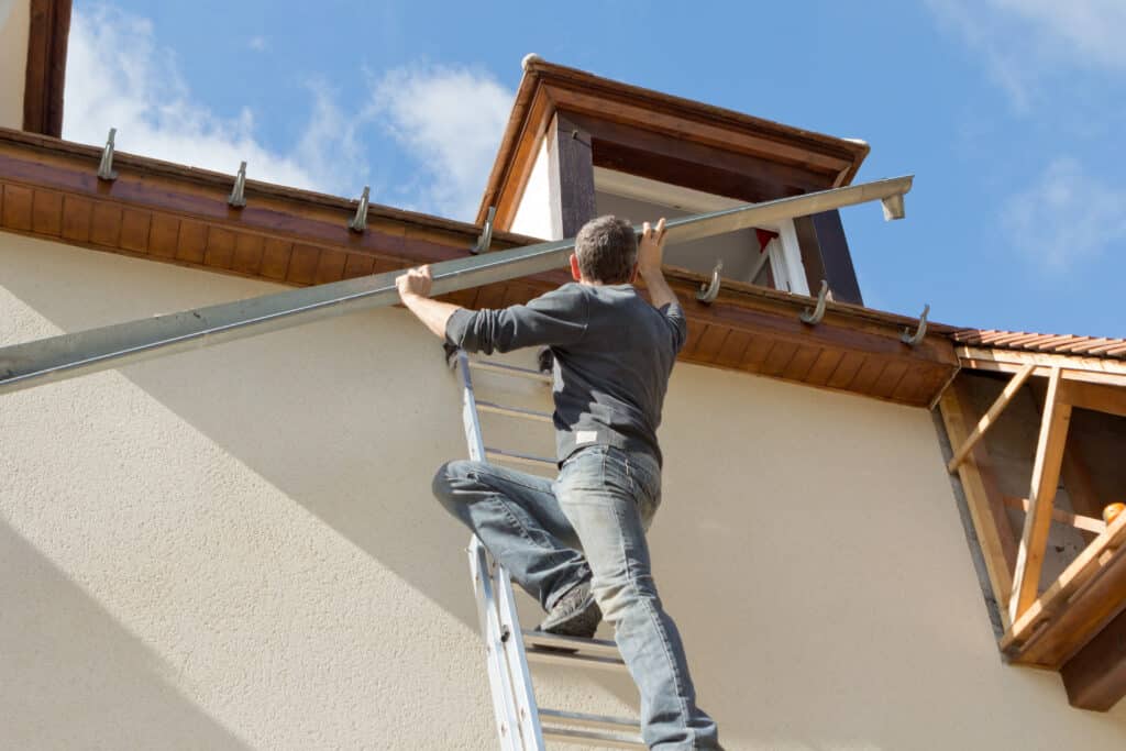 Homme sur échelle installe une gouttière métallique sur le toit d'une maison, ciel bleu nuageux en arrière-plan.