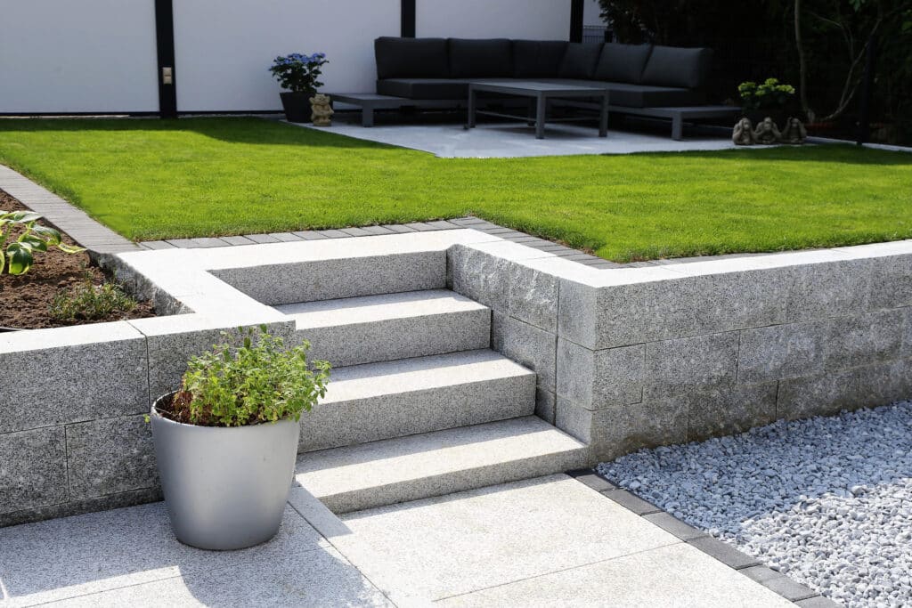 Aménagement de jardin moderne avec escalier et mur en granite, pelouse surélevée et patio carrelé.