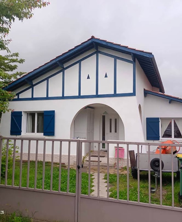 Charmante maison basque blanche avec boiseries bleues, volets bleus et toit de tuiles. Portail gris et jardin vert devant. Architecture régionale typique.