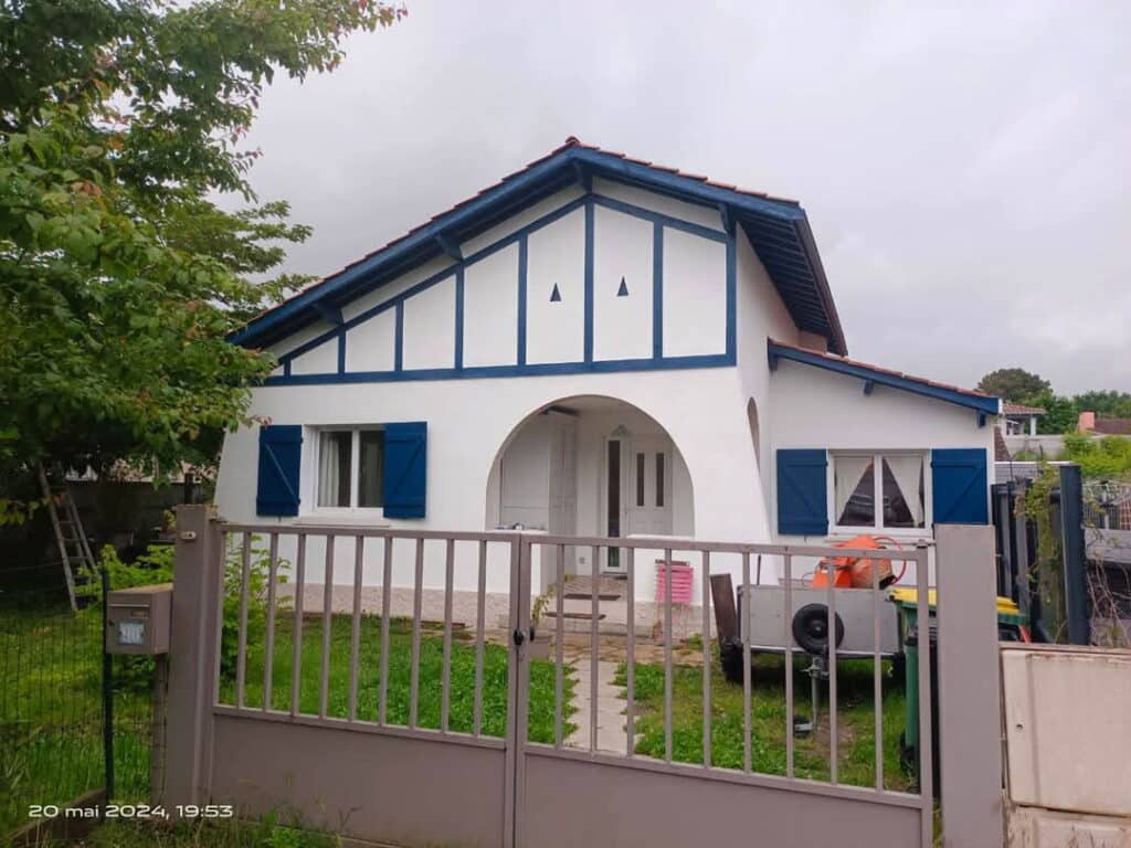 Maison basque blanche aux colombages et volets bleus. Portail gris, jardin, mixer, echelle. Date 20 mai 2024, 19:53.