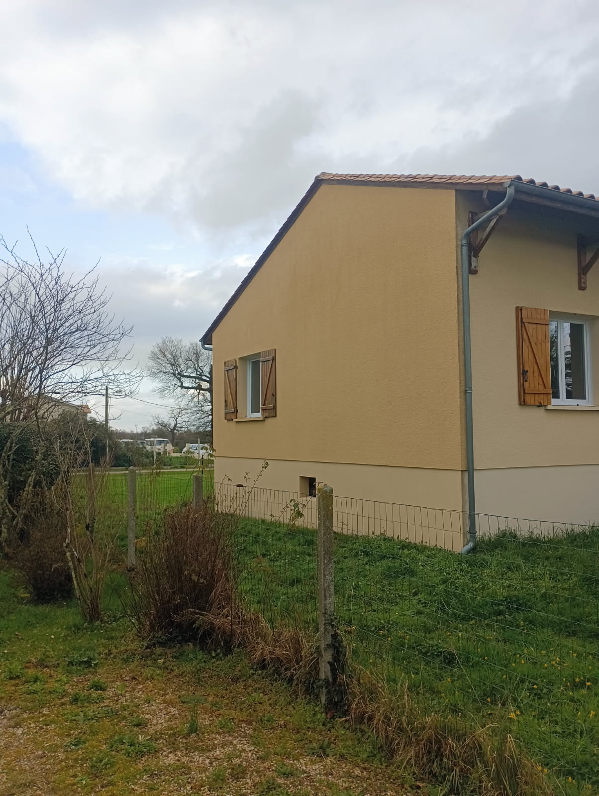 Maison à façade jaune et volets bois, avec clôture et jardin herbeux. Ciel nuageux.