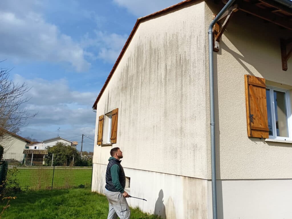 Homme pulvérisant un produit sur la façade d'une maison avec de larges traces sombres verticales. Nettoyage ou traitement anti-mousse.