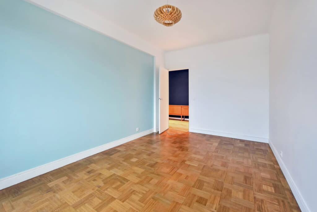 Pièce vide lumineuse, mur bleu ciel & parquet bois Pièce vide lumineuse murs bleu ciel et blancs, parquet bois motif tressé, porte ouverte sur espace sombre avec buffet. Luminaire design.