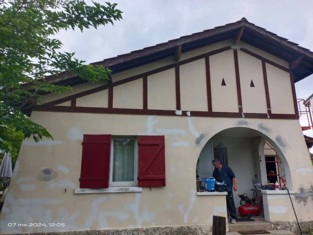 Rénovation façade maison à colombages: enduit appliqué, volets rouges, homme travaillant sous arche. 07 mai 2024.
