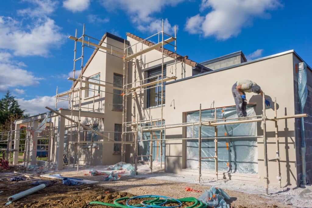 Ouvrier applique enduit sur façade beige de maison neuve sous ciel bleu nuageux. Échafaudages sur site de chantier.